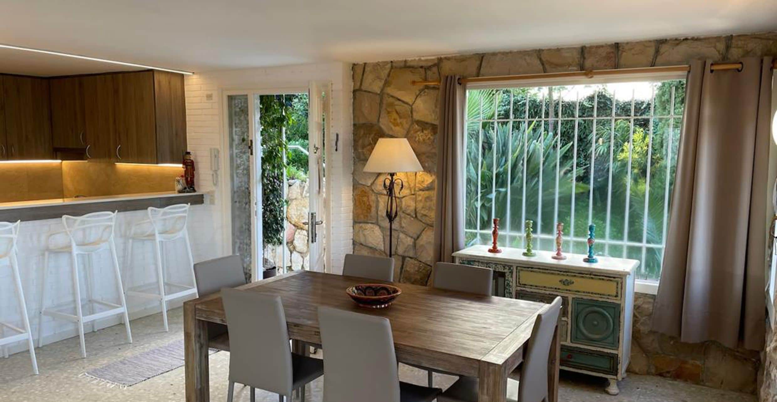 Een eetruimte met een stenen muur, een tafel met stoelen, een lamp en uitzicht op groen door een raam.