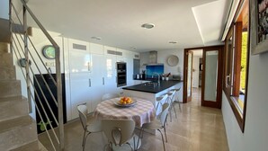 A modern kitchen with white cabinets, a central island with bar stools, and a dining table with a fruit bowl. - Villa in Uptown Pollensa with spectacular views and private pool (Pollença)