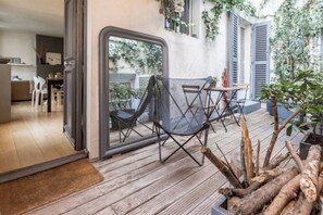 Een open woonoppervlak met een eettafel en stoelen, een zwarte klapstoel, en een houten terras met een plant in een pot.