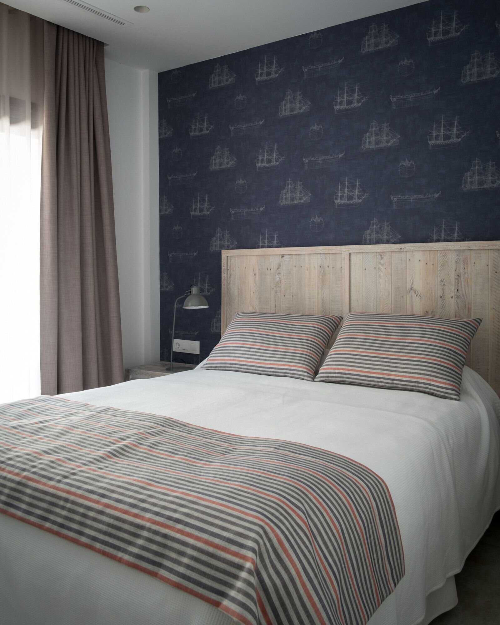 A neatly made bed with striped bedding, a wooden headboard, and a dark wall with ship patterns.