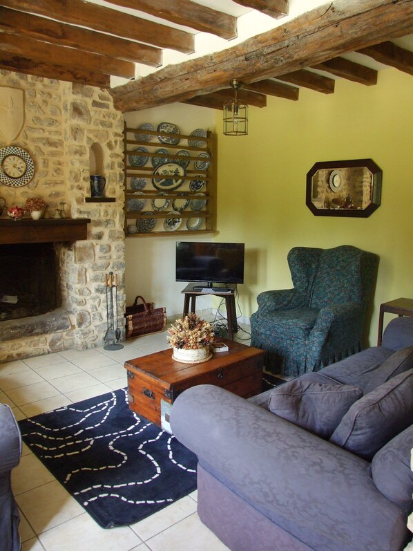 A cozy living room with a stone fireplace, a television, a patterned armchair, and a decorative wall shelf. - Traditional Normandy Cottage (La Bellière)