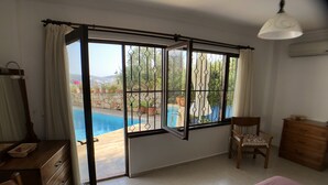 Ein Zimmer mit einem Bett, einem Sessel, einer Holzkommode, einem Schreibtisch mit Spiegel und einem Fenster mit Blick auf einen Swimmingpool und Grünflächen.