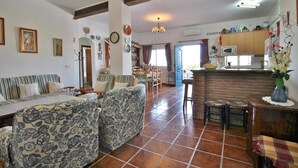 A cozy living room with patterned sofas, a dining area with chairs, and a kitchen with wooden cabinets and a tiled floor. - 1003 Villa Cornocalejo (Nerja)