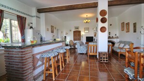 A spacious kitchen with a brick column, wooden stools, a dining area with a sofa, and a television. - 1003 Villa Cornocalejo (Nerja)