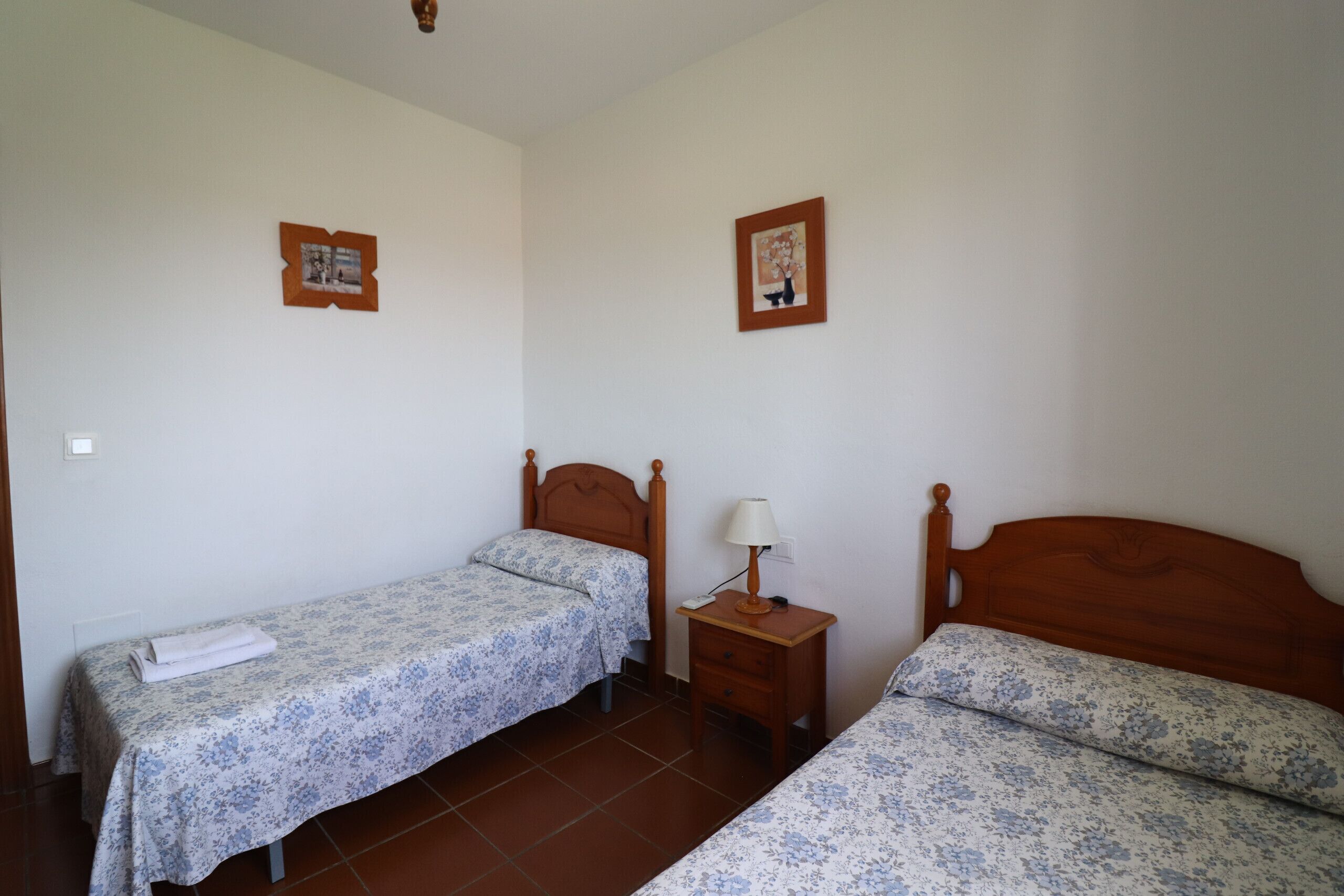 Une petite chambre propre, avec deux lits simples, une table de chevet avec une lampe et deux tableaux encadrés accrochés aux murs.