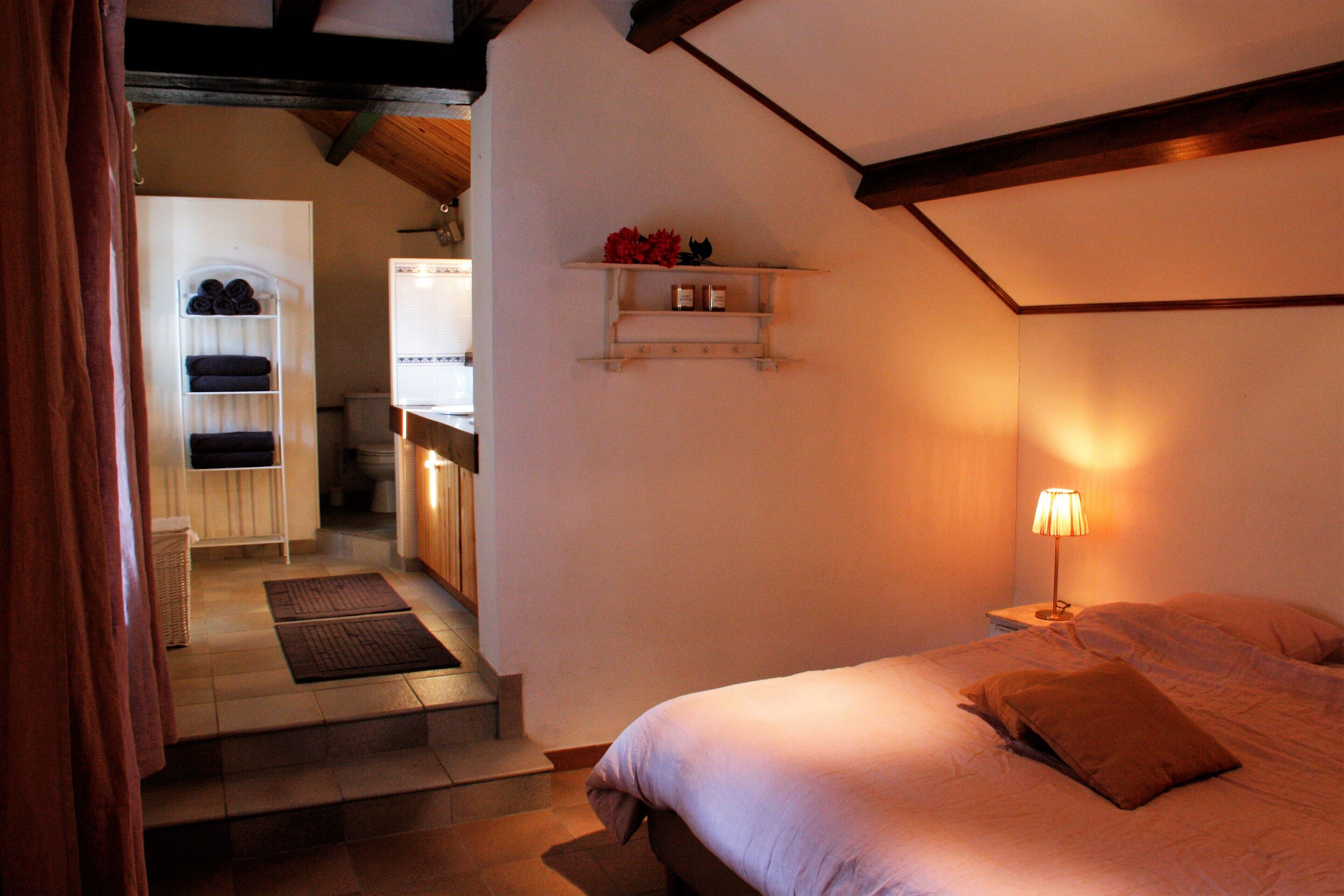 A bedroom with a bed, a nightstand, a lamp, a shelf with decorative items, and a bathroom with a towel rack and a basket.