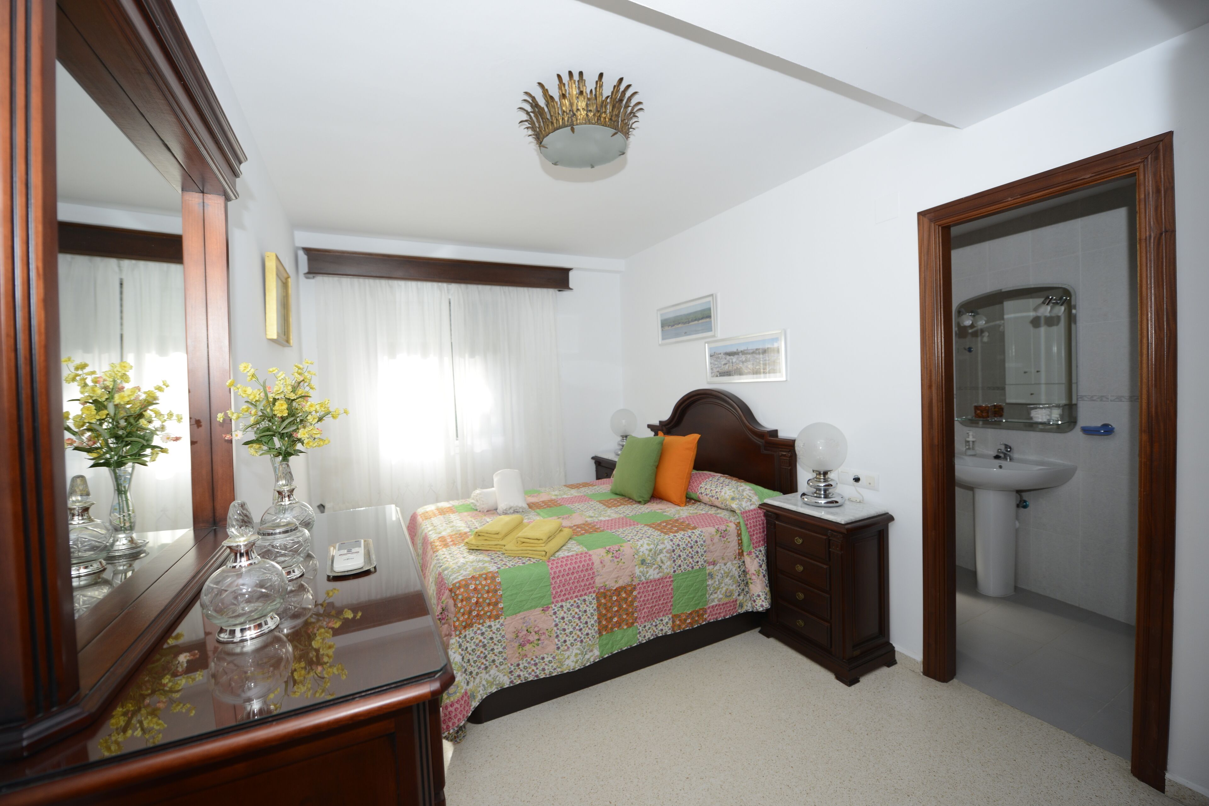 Een slaapkamer met een bed, nachtkastje, kaptafel en spiegel, plus een badkamer met een wastafel en spiegel.