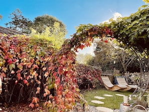 A garden with a pergola covered in ivy, a wooden deck with chairs, and a stone pathway.