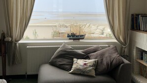 A living room with a large window, a sofa, a radiator, a wooden coffee table, and a bookshelf. - Beautiful 4* villa on the sand, feet in the water, stunning bay views (Le Crotoy)