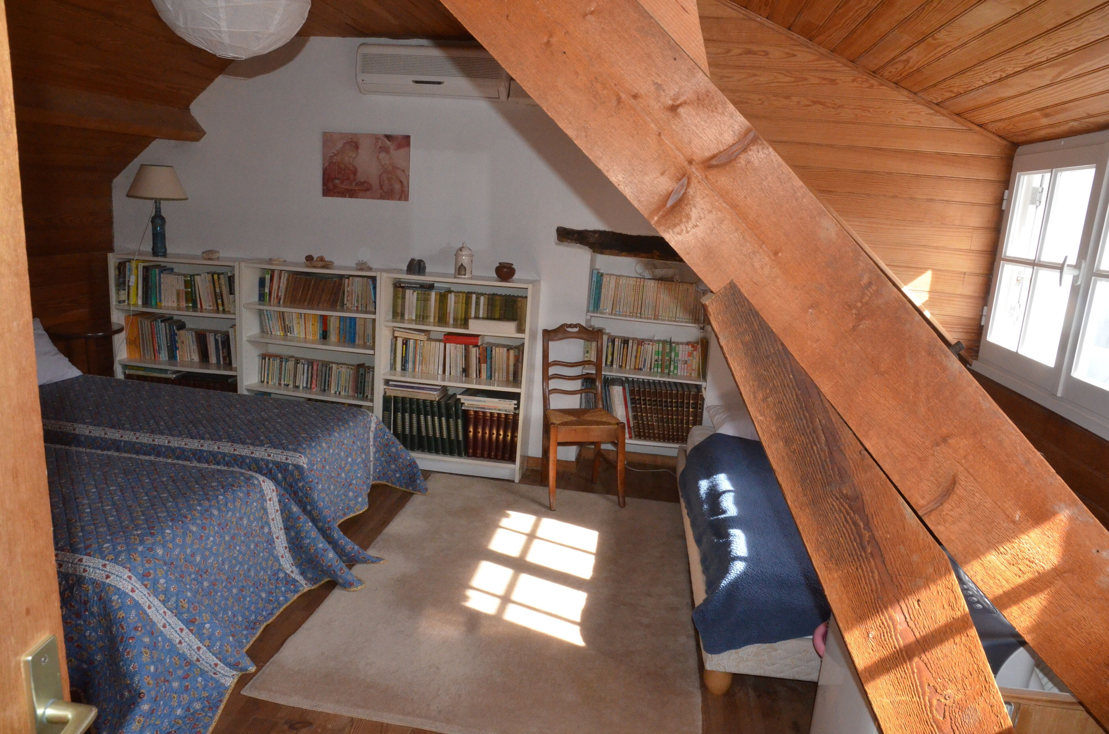 A cozy attic bedroom with a sloped ceiling, wooden beams, a large window, and a bookshelf filled with books.