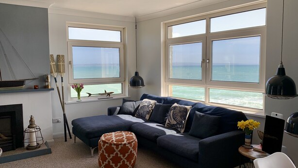 Un salon avec un canapé bleu foncé, un pouf à motifs et une vue sur la mer grâce à de grandes fenêtres.