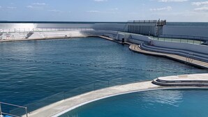 Une grande piscine à la forme incurvée, entourée d’une terrasse blanche et d’une rambarde en métal.
