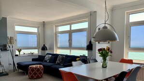 Un salon moderne avec un canapé, une table à manger et des chaises, de grandes fenêtres offrant une vue sur la mer, et un luminaire suspendu.