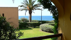 Uma varanda com vista para um jardim bem cuidado, uma palmeira e o mar.