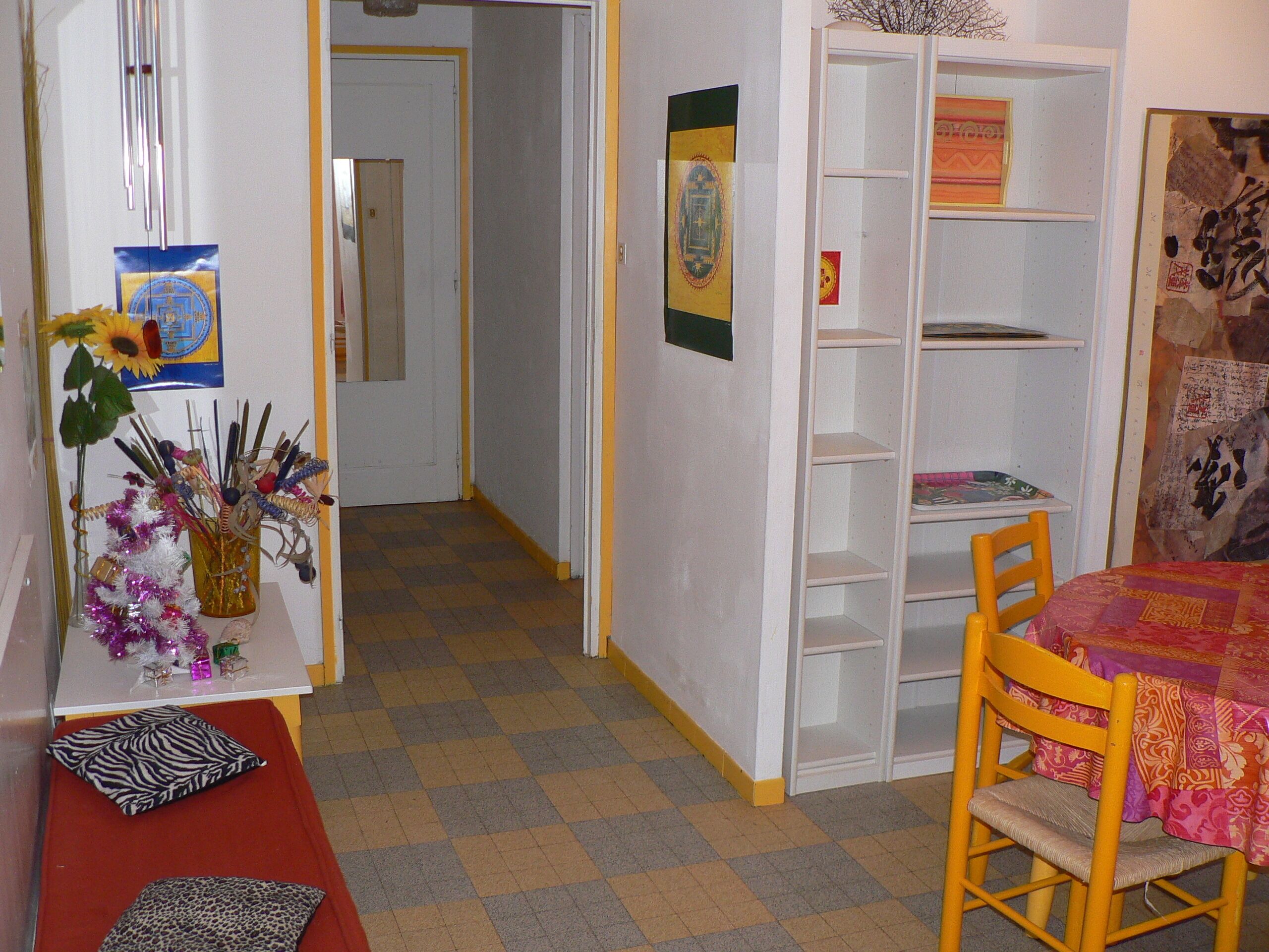 Ein Zimmer mit Schachbrettboden, einem Holzstuhl, einem Tisch mit farbenfroher Tischdecke, einem Bücherregal und einer Ziervase mit künstlichen Blumen.