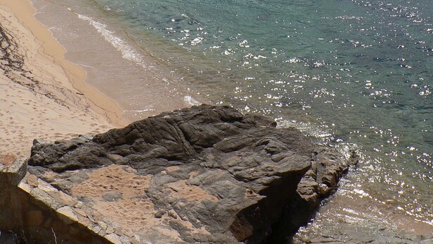 Uma praia com águas turquesa cristalinas, areal arenoso e formações rochosas.