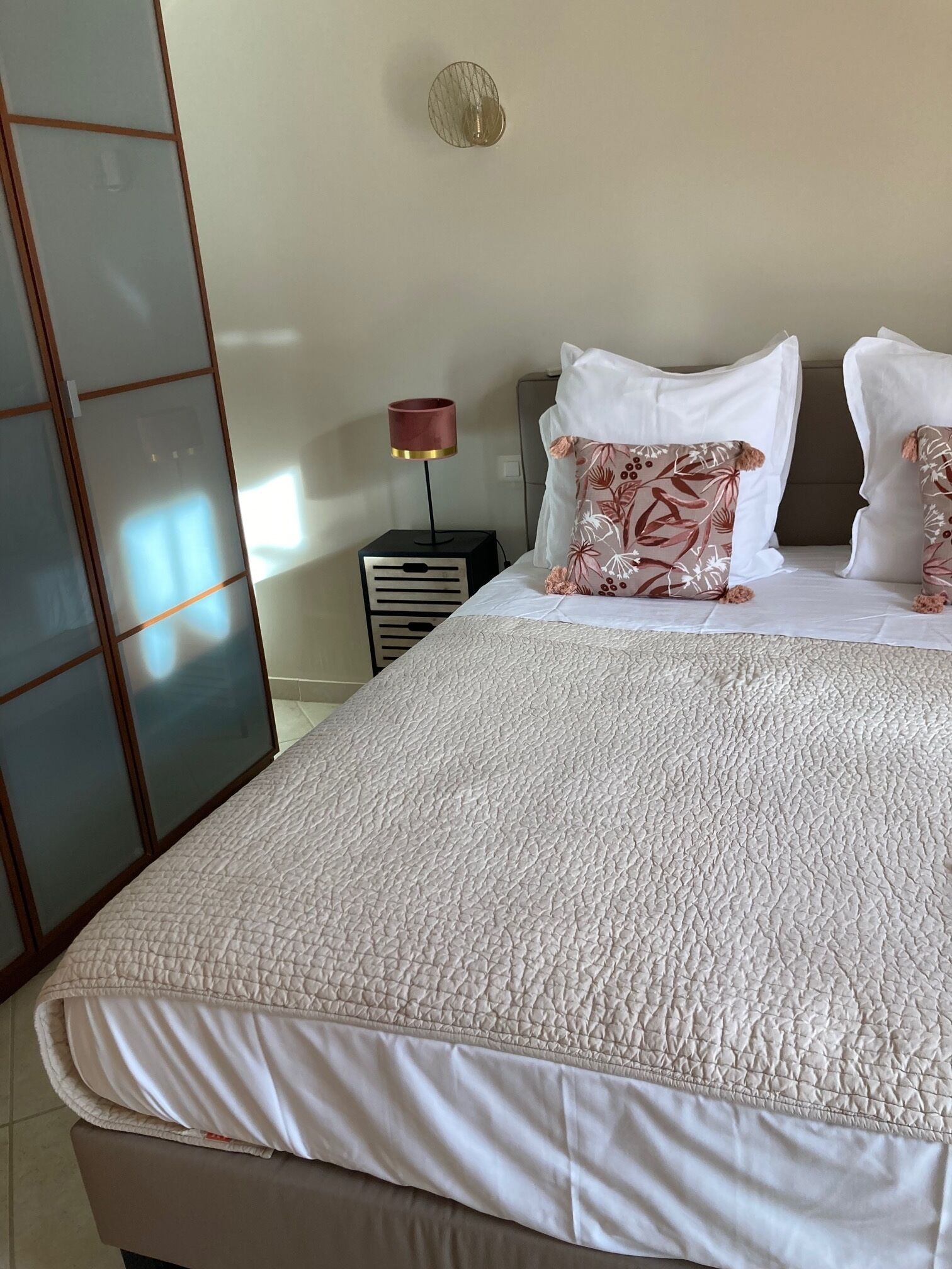 Un lit bien fait, recouvert d’une couette blanche à texture, avec deux oreillers blancs et un coussin à motifs floraux. Une petite table avec une lampe se trouve à côté du lit.