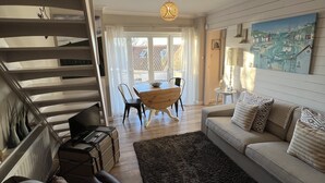 Een moderne woonkamer met een bank, een salontafel en een eettafel. Links is er een trap met een leuning en aan de muur hangt een schilderij.