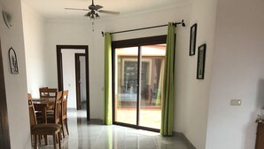 Un'ampia stanza con pavimento piastrellato, una ventola a soffitto, un'area pranzo con mobili in legno e una porta scorrevole in vetro che dà su uno spazio esterno.