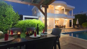 Un espace de restauration en plein air avec une table dressée pour deux personnes, entouré d’une végétation luxuriante et d’une piscine.