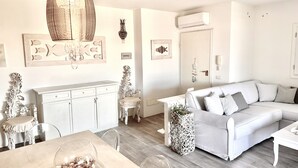 A modern living room with a white sofa, a dining table with a glass bowl, and a decorative chandelier. - House 50 meters from the sea residence S'ABBA E SA PEDRA  (Golfo Aranci)