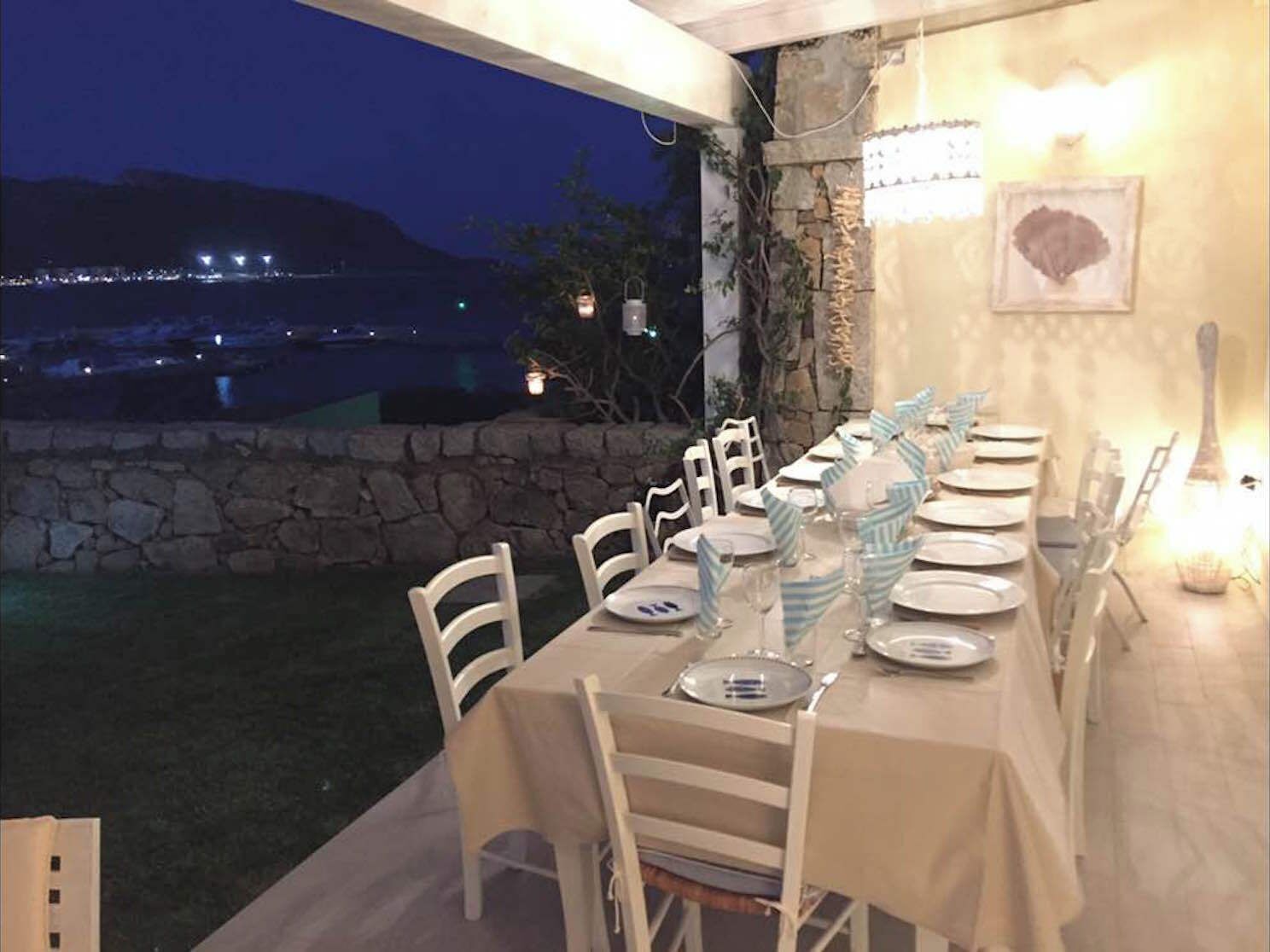 An outdoor dining area with a stone wall and a view of the sea at night.