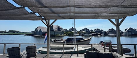 Un espace extérieur abrité, aménagé avec des sièges, offrant une vue sur un port de plaisance et les maisons situées de l’autre côté de l’eau.