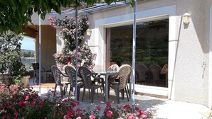 Une terrasse extérieure avec une table et des chaises, entourée de fleurs roses et d’un rosier grimpant.