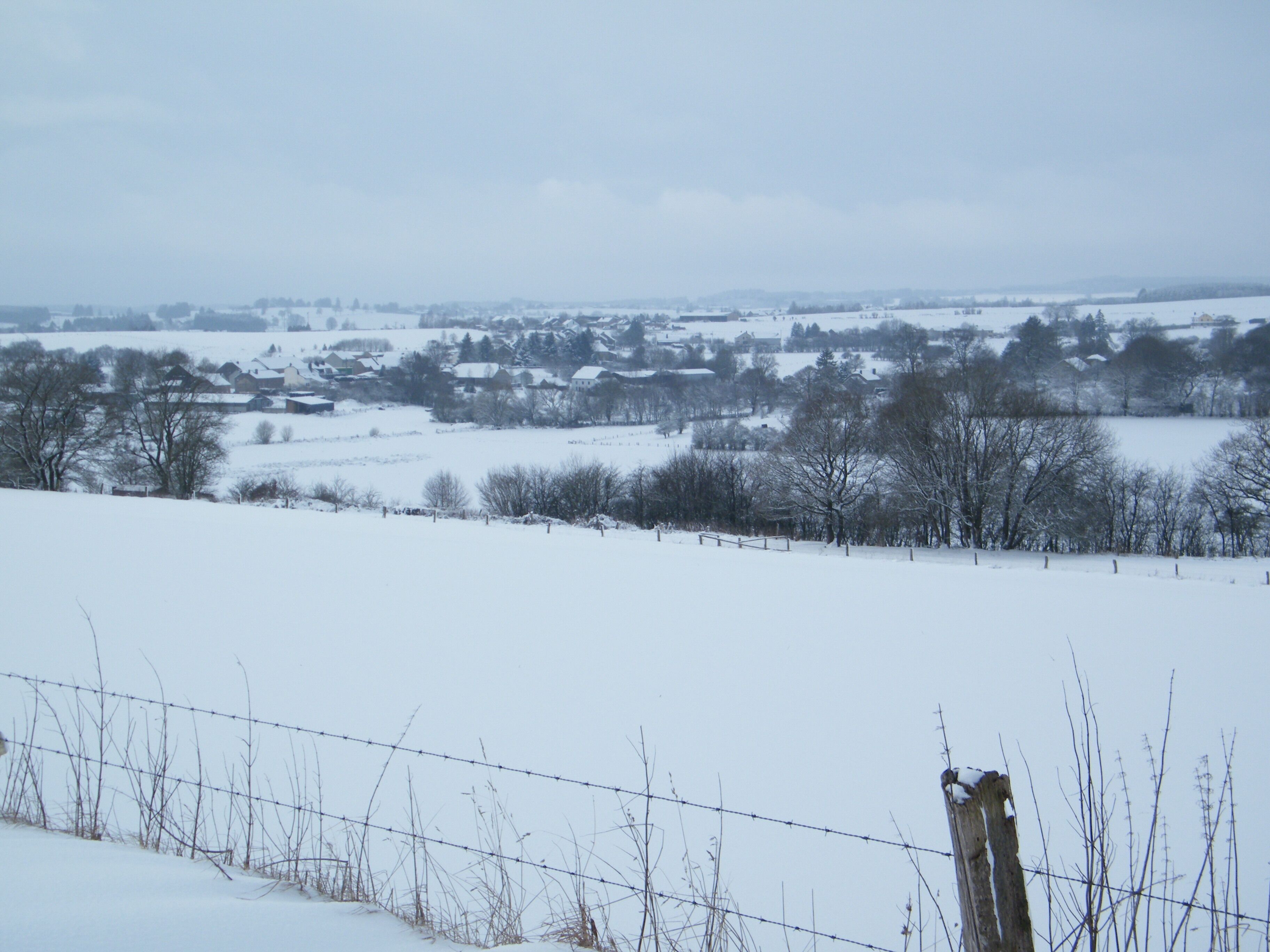 Schneebedeckte Landschaft mit einem Stacheldrahtzaun im Vordergrund und einem Dorf in der Ferne.