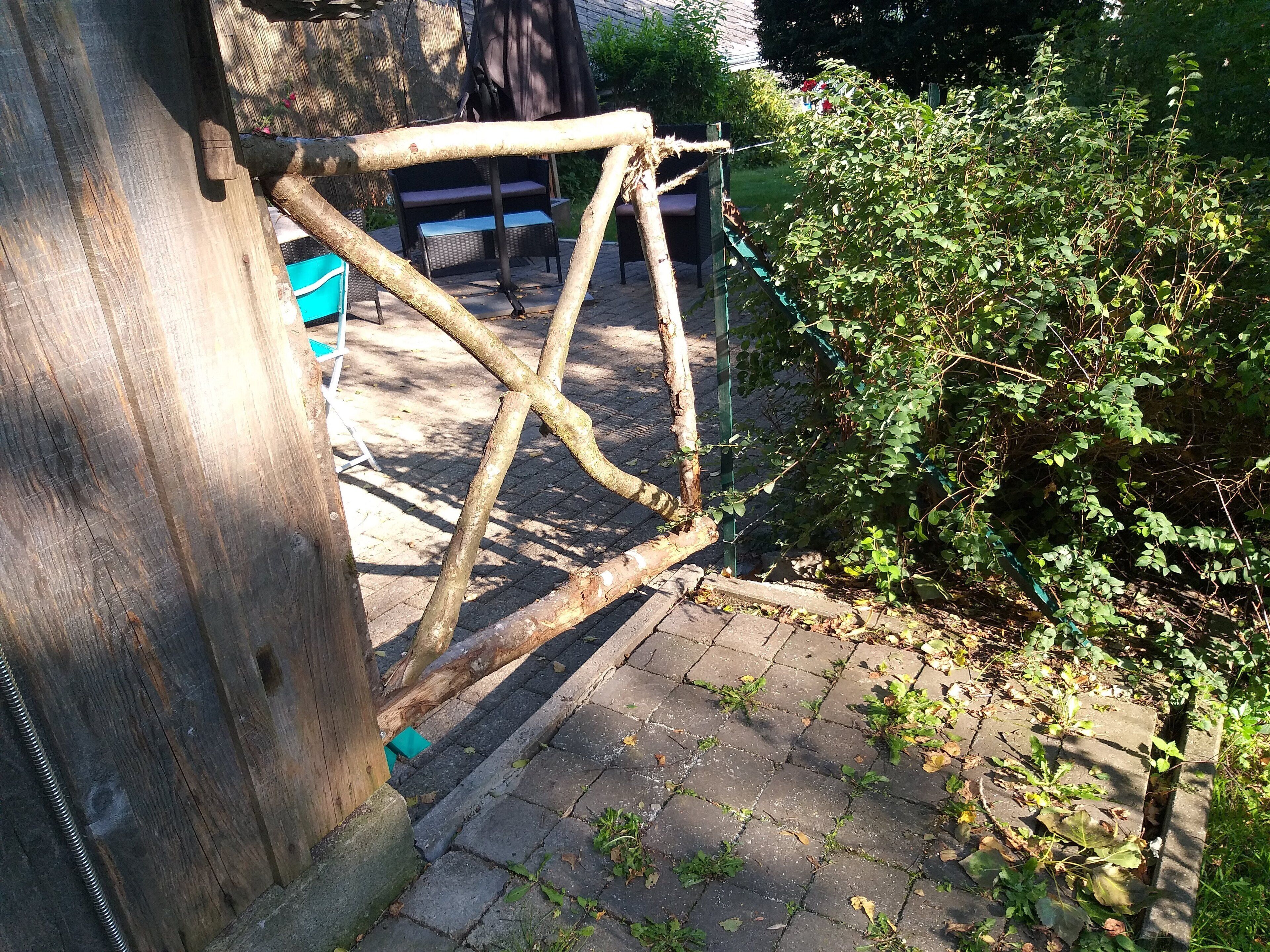 A rustic wooden structure with a metal frame, possibly a garden feature or play equipment, adjacent to a paved walkway with a green bush.