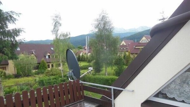 Un balcón con un banco de madera, una antena parabólica y vista a casas y montañas al fondo.