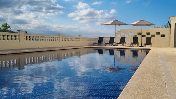 En swimmingpool med liggestole og parasoller, omgivet af en lav mur og en klar himmel.