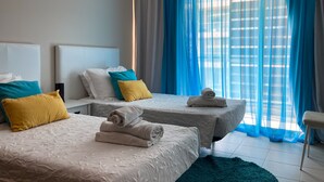 Ein Hotelzimmer mit zwei Betten, jeweils mit weißer Bettdecke und einem zusammengerollten Handtuch. Blaue und gelbe Kissen, und ein großes Fenster mit blauen Vorhängen.