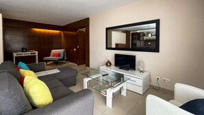 Ein modernes Wohnzimmer mit einem grauen Sofa, einem Glastisch, einer weißen TV-Konsole und einem großen Spiegel an der Wand.