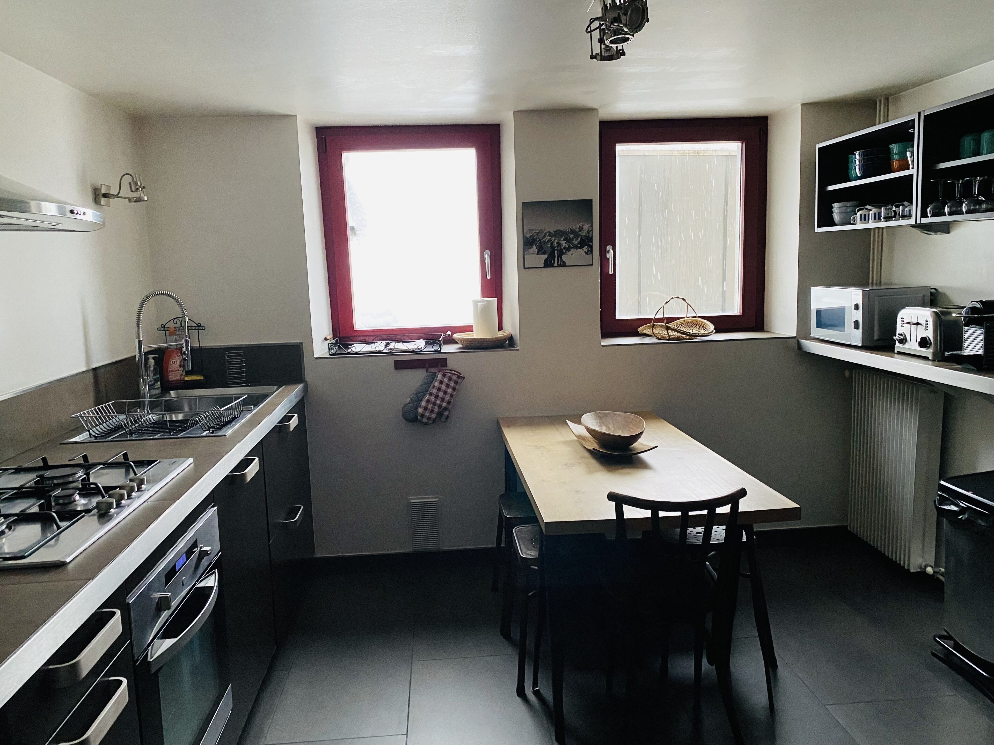 A compact kitchen with a gas stove, oven, and microwave. There is a window with a red frame and a small table with a bowl.