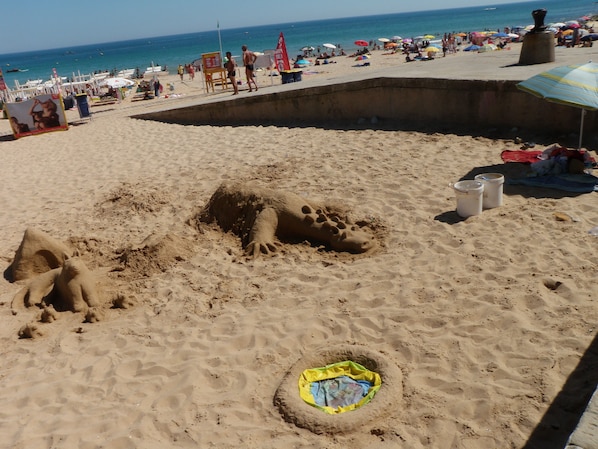 Une plage de sable où se trouve une grande sculpture de dinosaure, ainsi qu’un petit trou contenant un objet coloré.