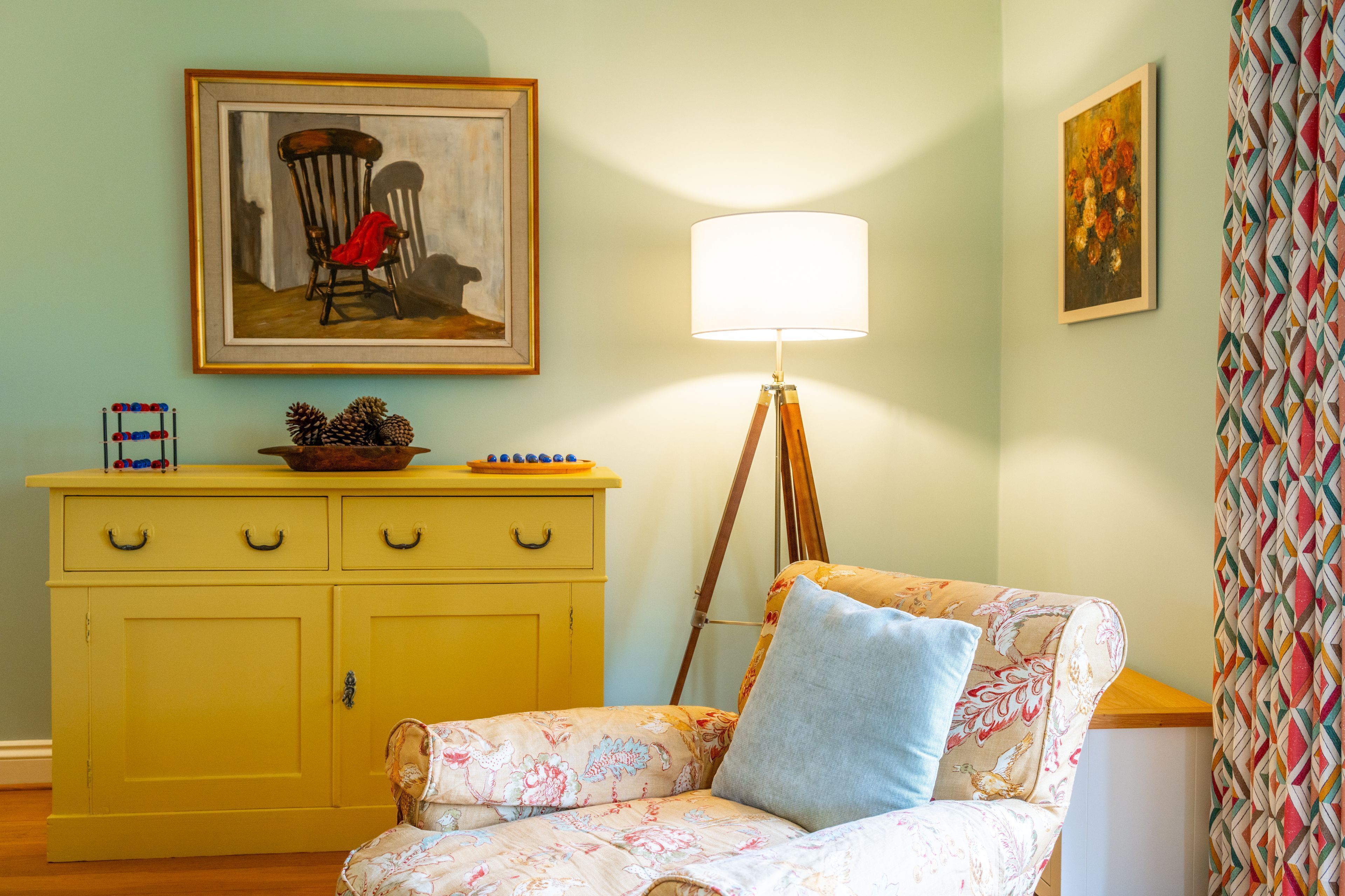 Una habitación con un aparador amarillo, una silla con tapicería floral, una lámpara de pie y dos obras de arte enmarcadas en las paredes.