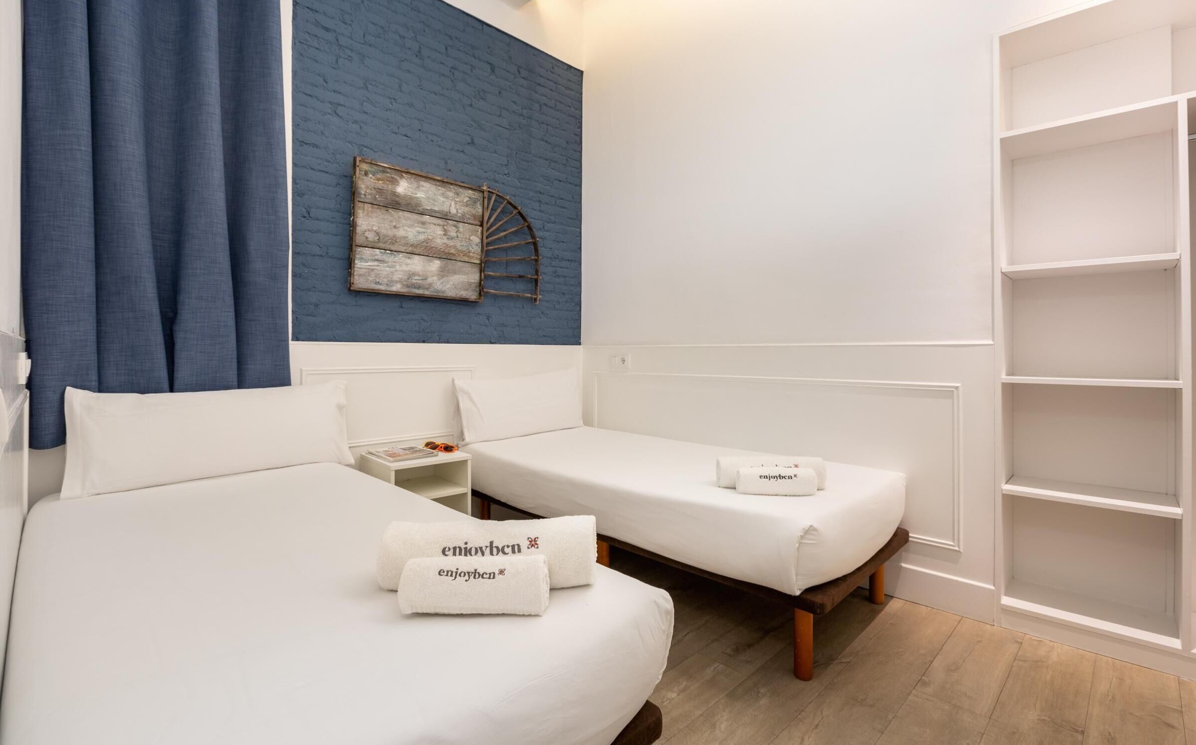 Habitación de hotel con dos camas individuales, una pared decorativa de ladrillo azul, un armario blanco y una mesita de noche pequeña con una revista y zapatillas.