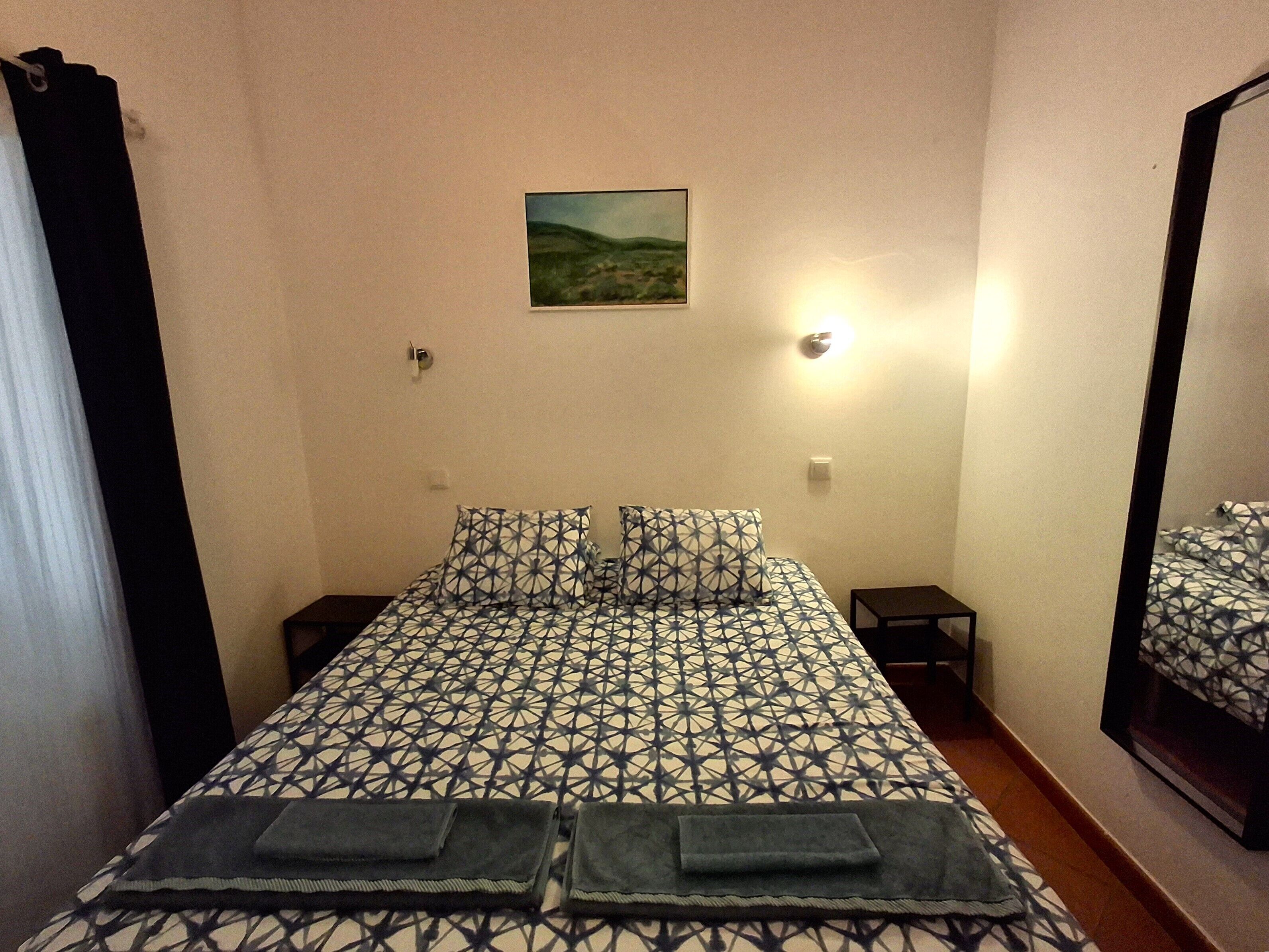 A bedroom with a patterned bedspread, two pillows, a small bedside table, a nightstand, a wall-mounted light, and a painting above the bed.