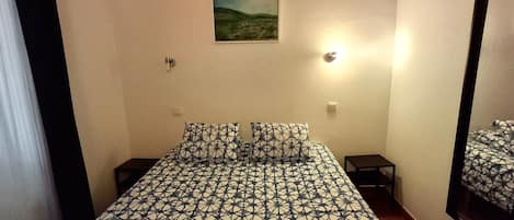 A bedroom with a patterned bedspread, two pillows, a small bedside table, a nightstand, a wall-mounted light, and a painting above the bed.