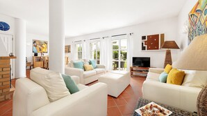 A spacious living room with white sofas, a coffee table, and a TV. There are large windows with curtains, a brick wall, and a painting on the wall. - Villa Farol OCV - Ocean Front (Galé)