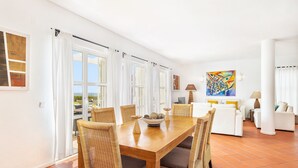 A spacious living area with a wooden dining table, wicker chairs, and a view of the outdoors through large windows. - Villa Farol OCV - Ocean Front (Galé)