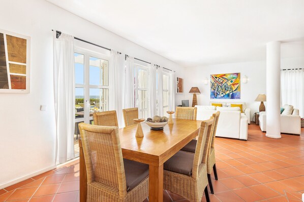 A spacious living area with a wooden dining table, wicker chairs, and a view of the outdoors through large windows.