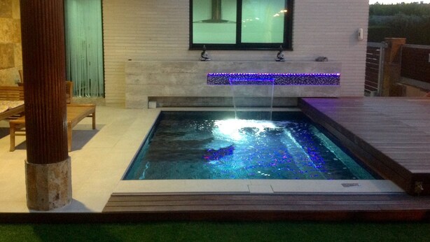 Piscina rectangular con caída de agua, rodeada por una terraza de madera y un edificio blanco con una ventana.