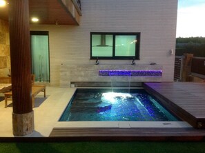 Une piscine rectangulaire agrémentée d’une cascade, entourée d’une terrasse en bois et bordée d’un bâtiment blanc doté d’une fenêtre.