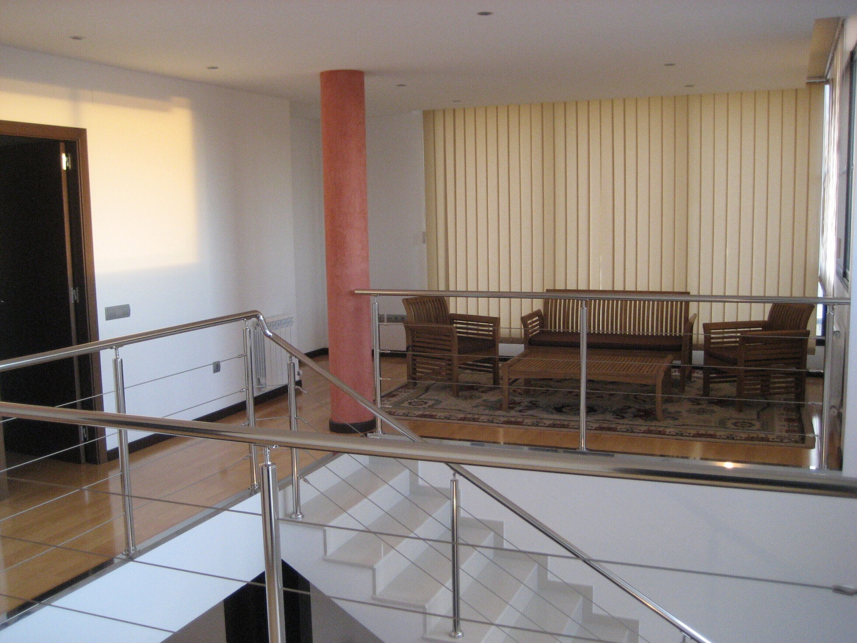 A spacious living area with a high ceiling, a staircase with a metal railing, and a large window with vertical blinds.