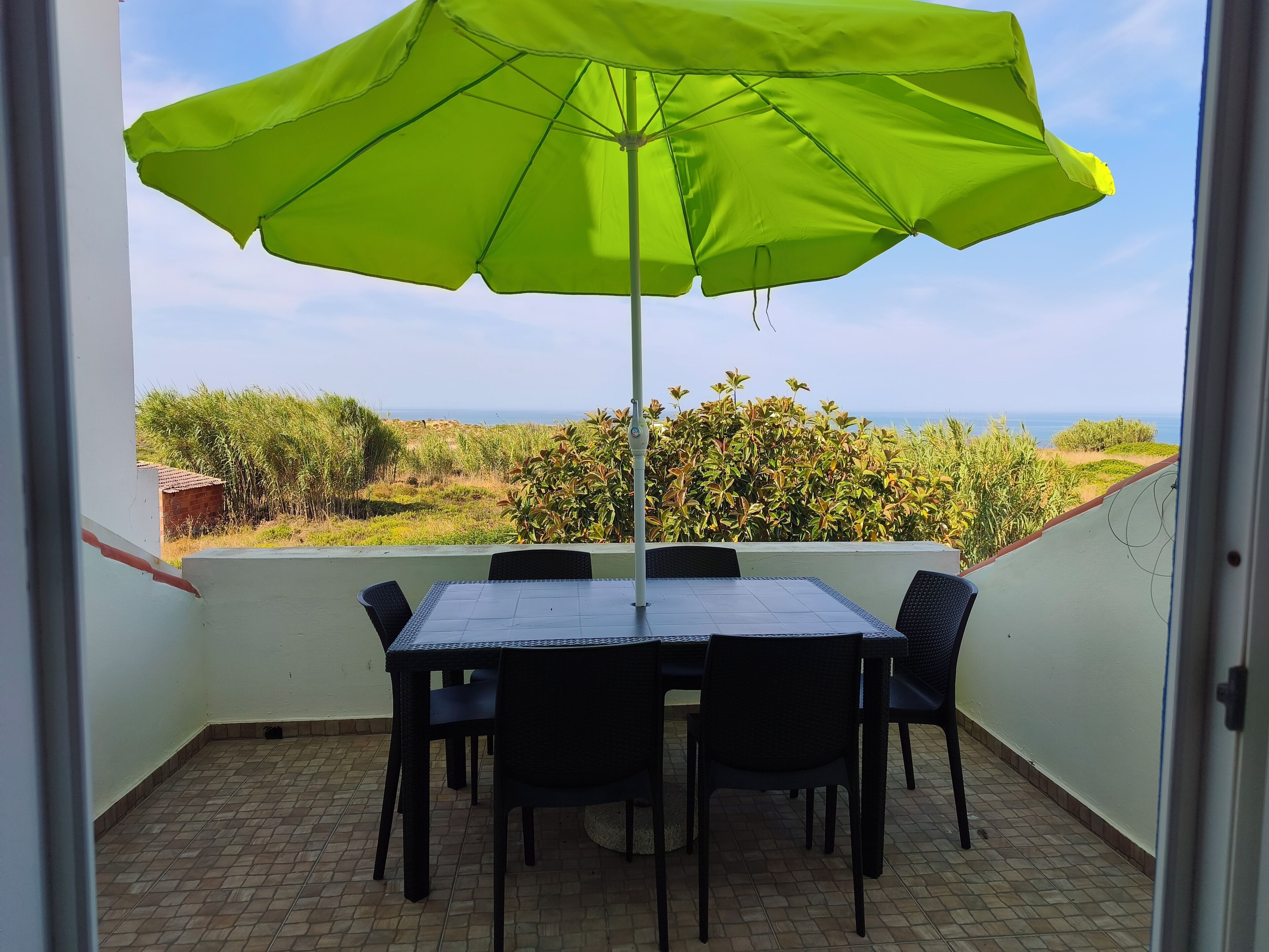 Ein Balkon mit Tisch und Stühlen, einem grünen Sonnenschirm und Blick aufs Meer.