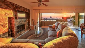 A living room with a stone fireplace, a ceiling fan, a large sofa, and a view of the sunset through the balcony doors. - Villa Callista -  Modern Villa that sleeps 8 guests  in 4 bedrooms (Paphos)