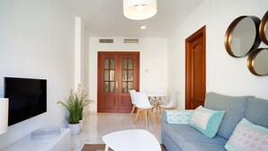 A modern living room with a flat-screen TV, a white coffee table, a blue sofa with patterned cushions, a dining table with chairs, and a wooden door with glass panels. - iloftmalaga Puerta del Mar II (Málaga)