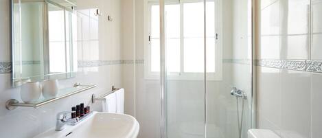 Un baño moderno con ducha envuelta en vidrio, un lavamanos blanco con espejo y una pequeña repisa con artículos de aseo.